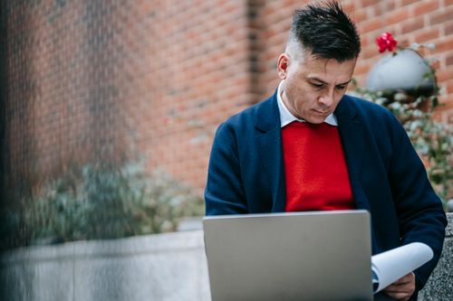 Free Adult man in formal attire working remotely on a laptop outdoors against a brick wall. Stock Photo