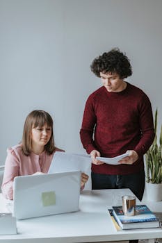 Two colleagues working together in a modern office setting with laptops and documents.