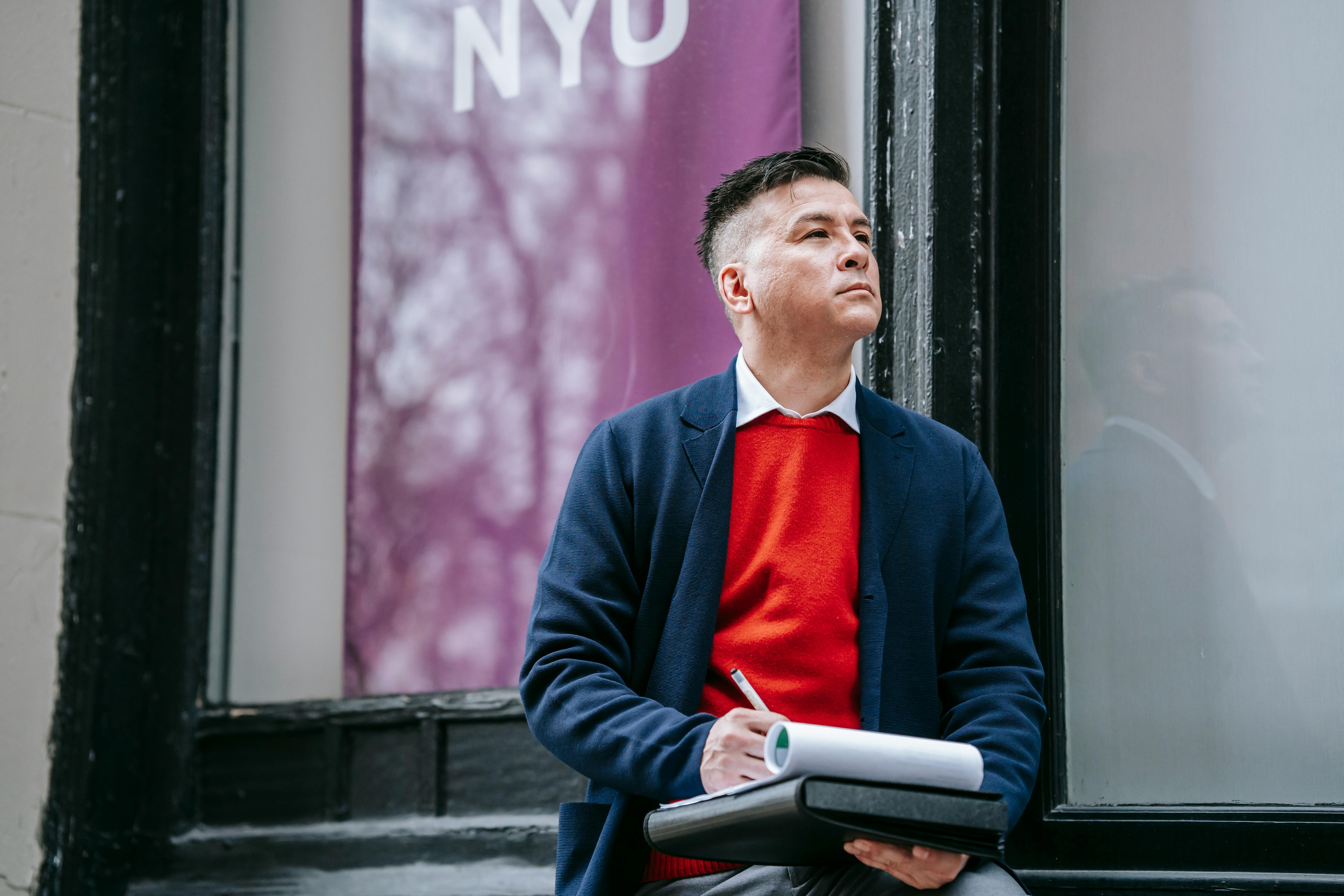 Professional man in a red sweater writing outside NYU. Urban academic setting.