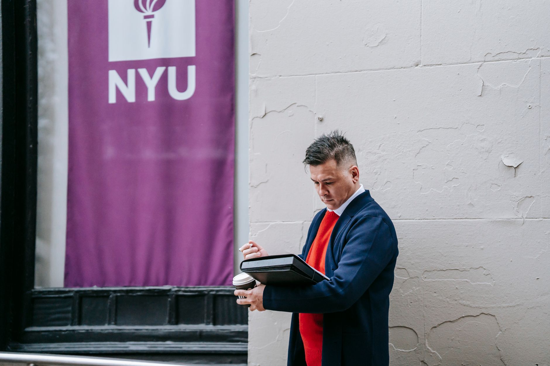 A man in business attire standing outside near an NYU banner, working on a laptop.