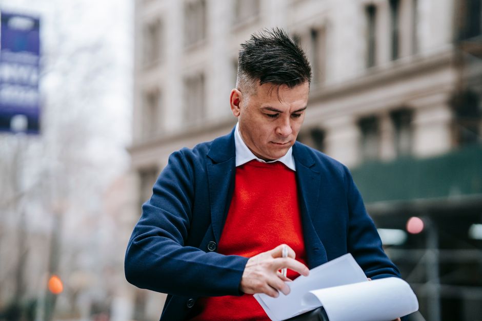 Man in a stylish outfit examines paperwork outside a city building, focused and pensive.