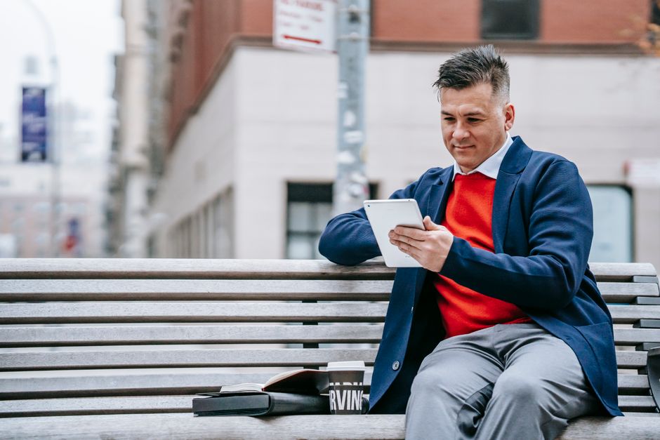 Businessman in stylish attire using a digital tablet outdoors, relaxing on a wooden bench. Businessman in stylish attire using a digital tablet outdoors, relaxing on a wooden bench.