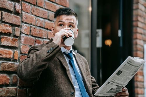 Free Dapper man in a formal coat drinks coffee while reading a newspaper outdoors. Stock Photo