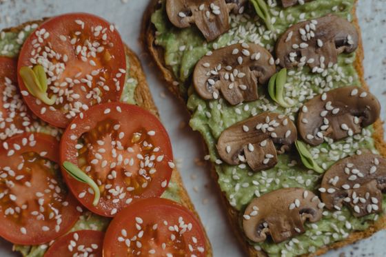 Two toasts with mashed avocado, one topped with tomato slices and one with mushrooms, sesame seeds