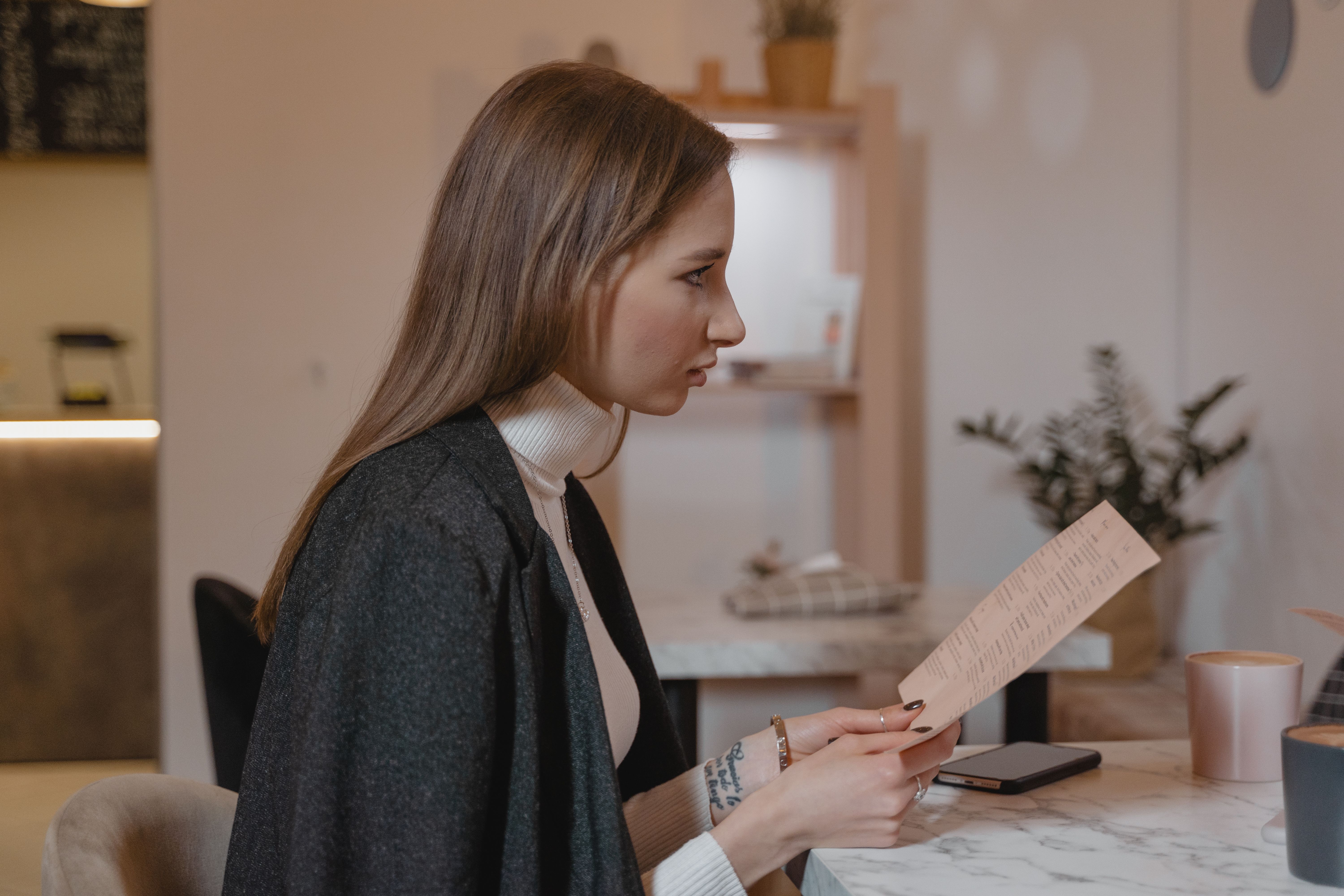 Side view of woman holding menu in a cozy caf&eacute; setting, wearing a turtleneck and jacket.