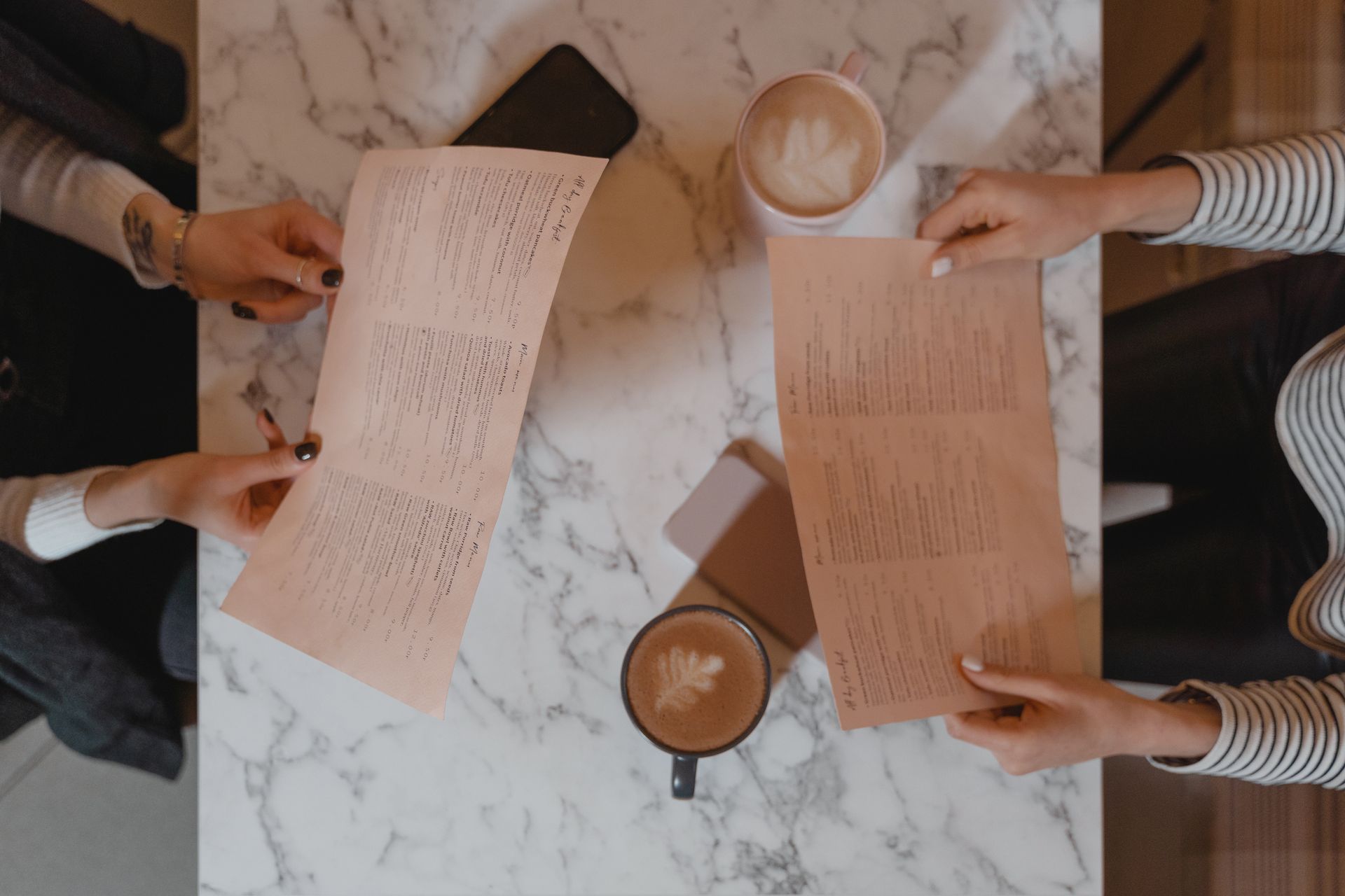 Overhead view of two friends sitting at a marble table, holding menus and enjoying coffee.