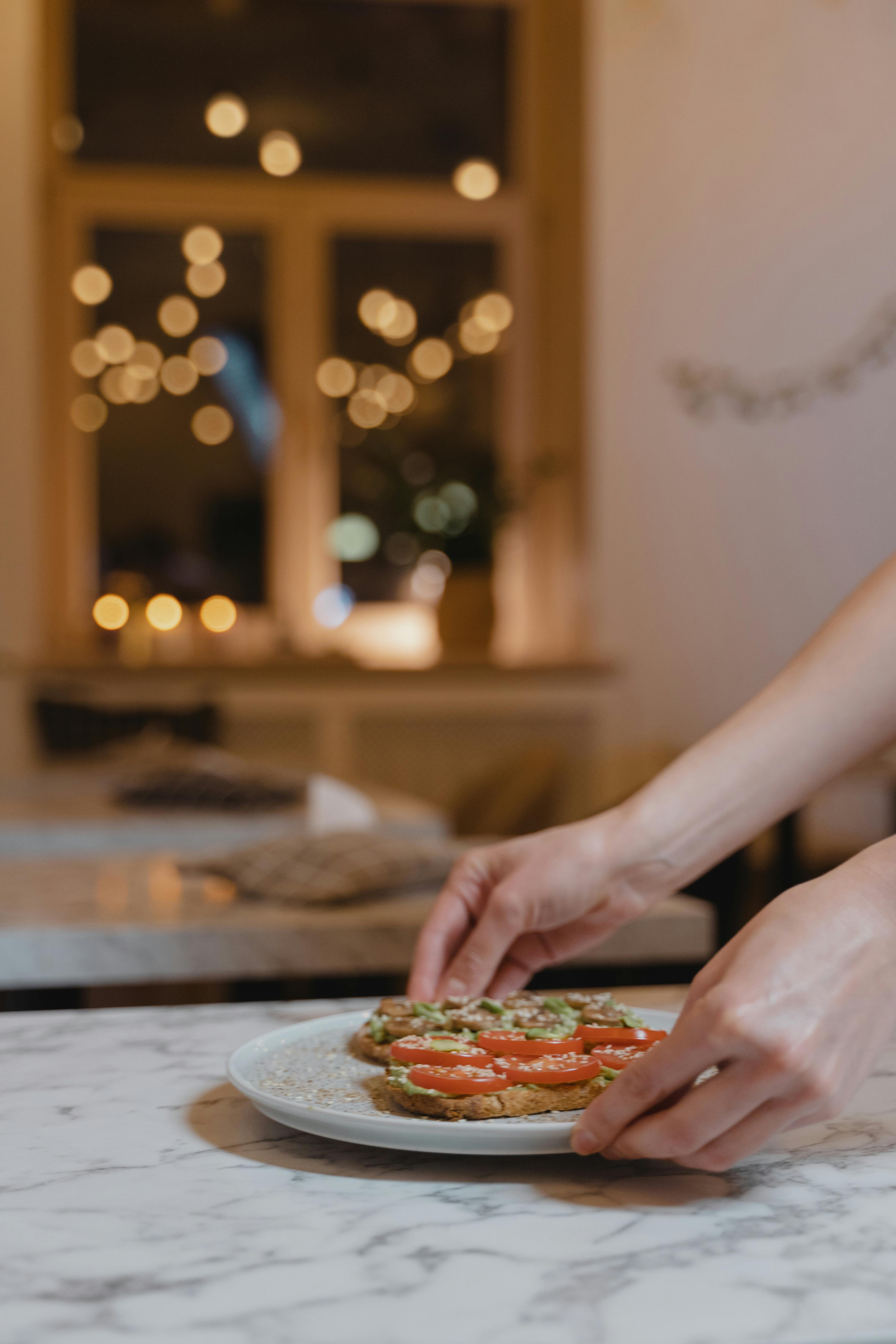 Close-up of hands adjusting avocado toast with tomato slices on a ceramic plate indoors.