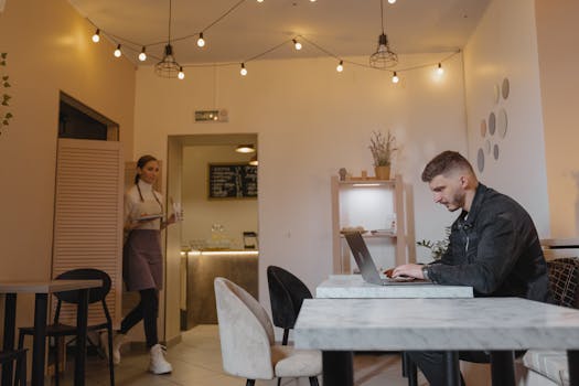 Man working on laptop in a stylish café while a waitress serves customers.