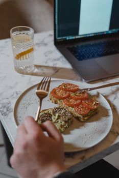 A delicious vegan brunch setup with avocado toast, tomatoes, and a refreshing drink beside a laptop.