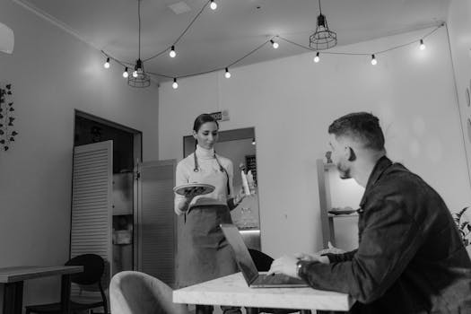 Grayscale image of a café scene with a waitress serving a customer working on a laptop.