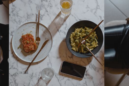Overhead view of a vegan pasta and tomato salad meal on a marble table with a phone.