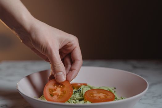 A close-up photo of a hand placing fresh tomato slices on a vegetable salad in a bowl.