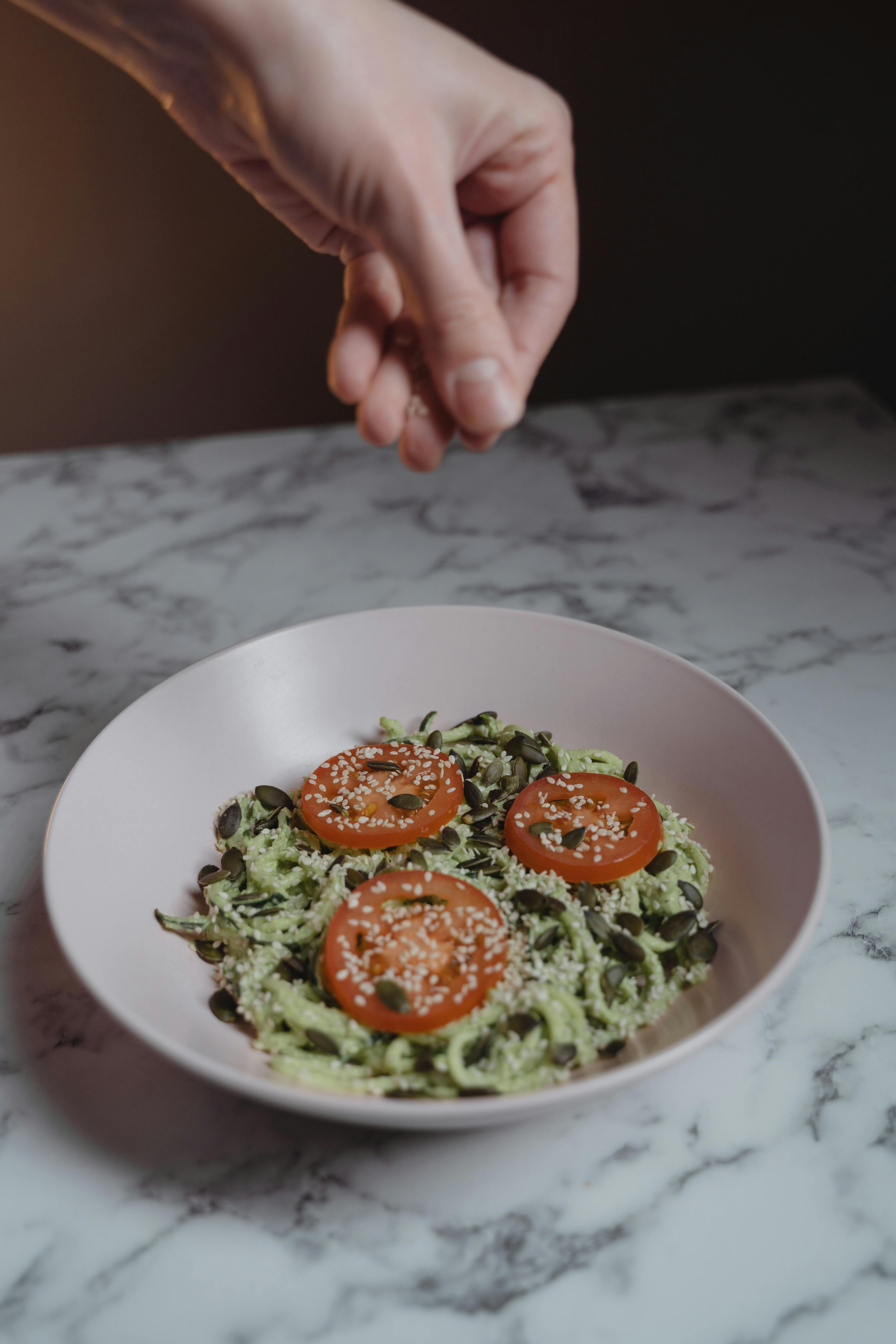 Hand sprinkling sesame seeds on fresh tomato pasta over a marble table.