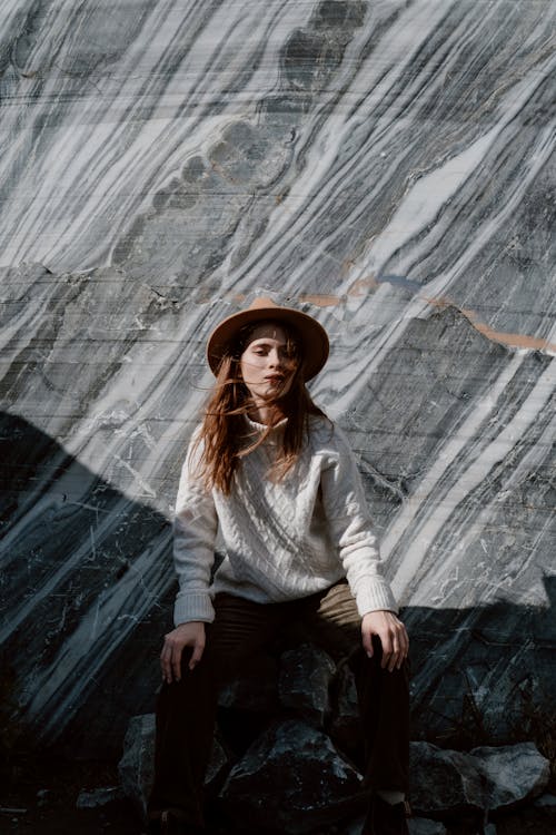 Woman in White Long Sleeve Shirt and Black Pants Wearing Brown Hat Sitting on Rock