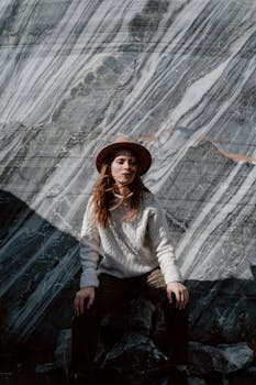 A woman in a hat and sweater sits by a marble rock formation, basking in nature.