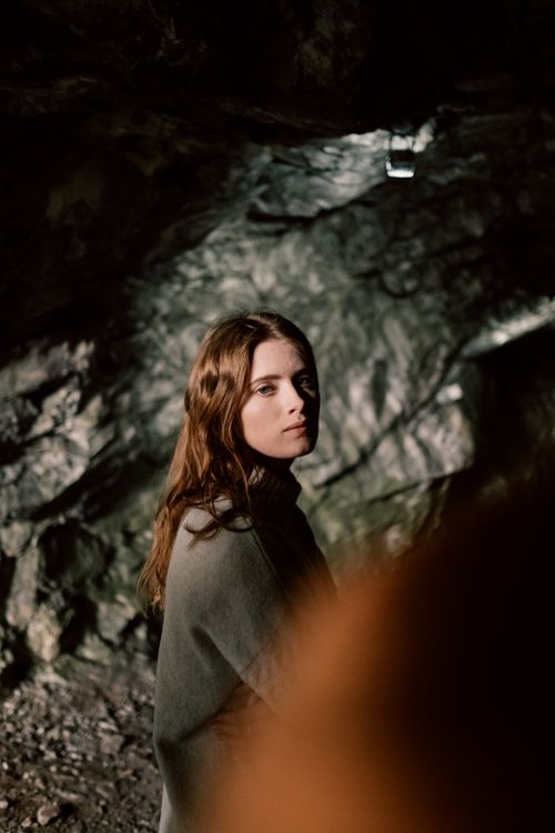 Free A woman with long hair and gray clothing stands thoughtfully in a dimly lit cave. Stock Photo