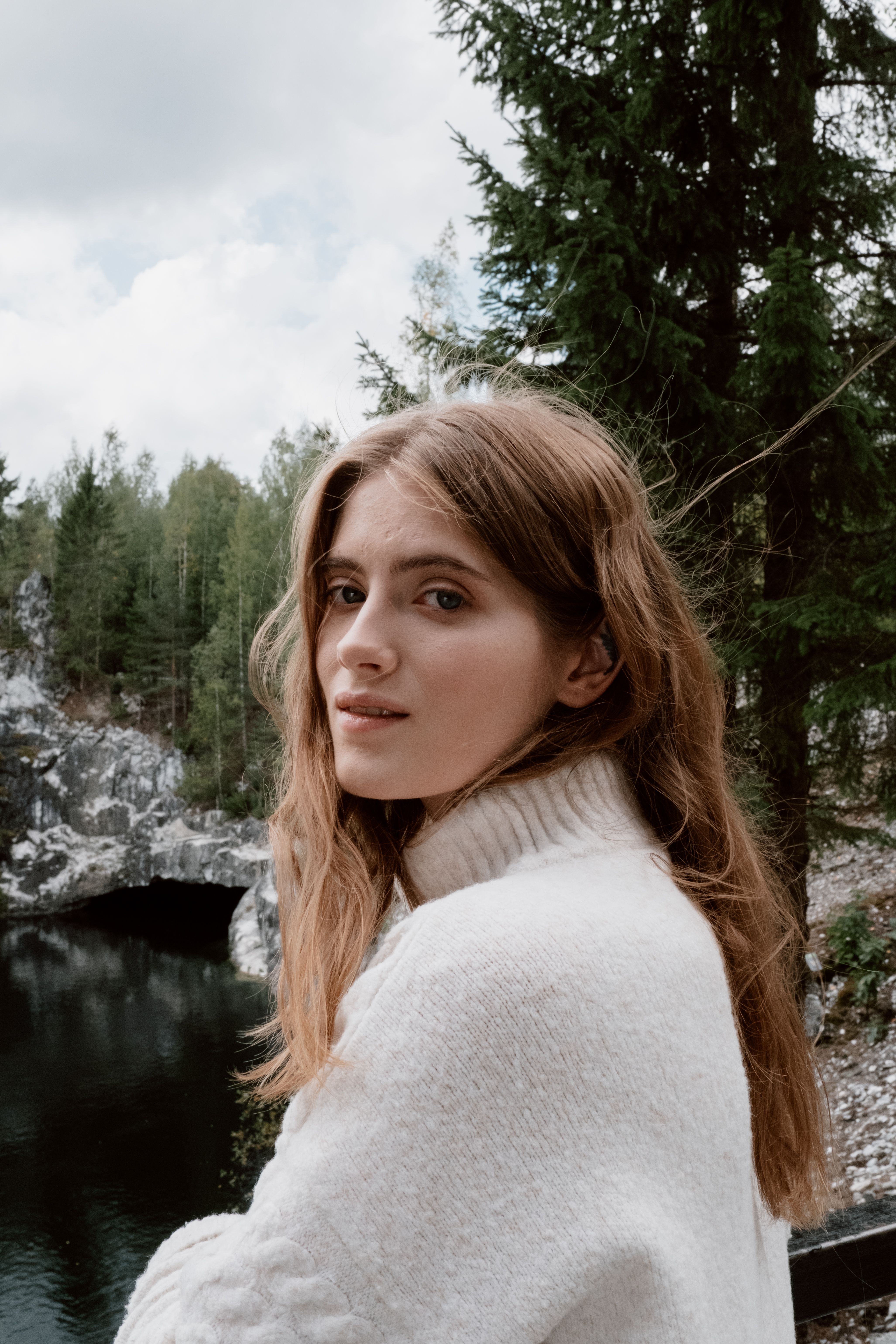 A serene brunette woman in a white sweater stands by a scenic river, surrounded by lush greenery.