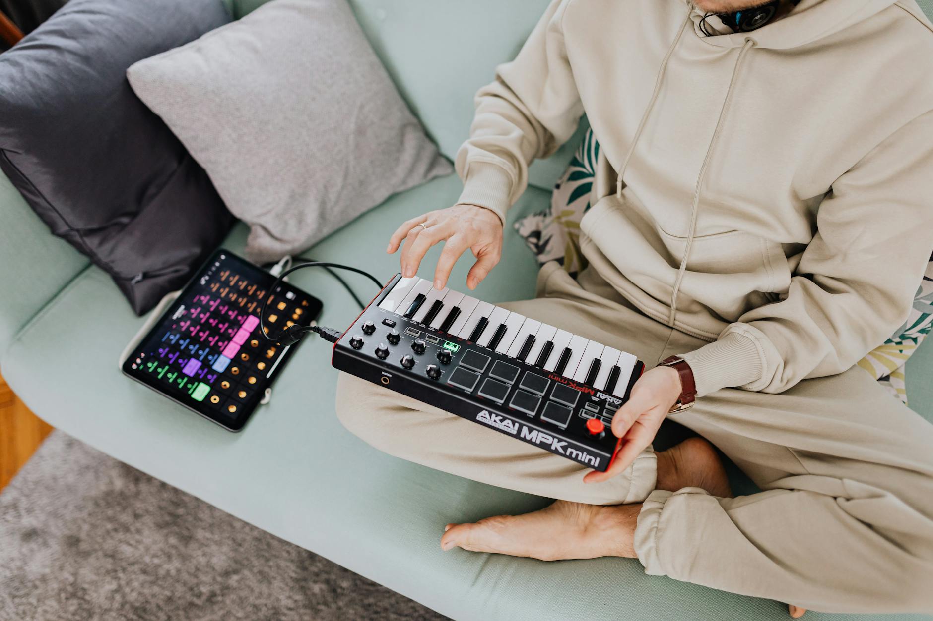 A person in a beige tracksuit using Akai MPK Mini controller and a tablet on a sofa.