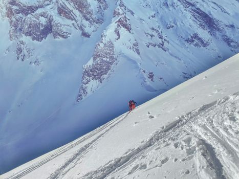 Unrecognizable skiers skiing on snowy vast valley towards rocky severe mountain range on sunny winter day