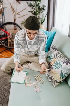 A young man wearing a beanie counts cash while sitting on a couch, surrounded by notes and a notebook.