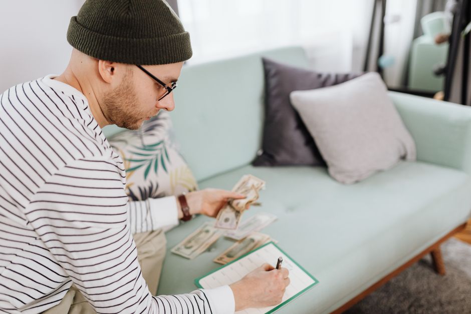 Adult man with beanie counting cash and making notes on a couch. Adult man with beanie counting cash and making notes on a couch.