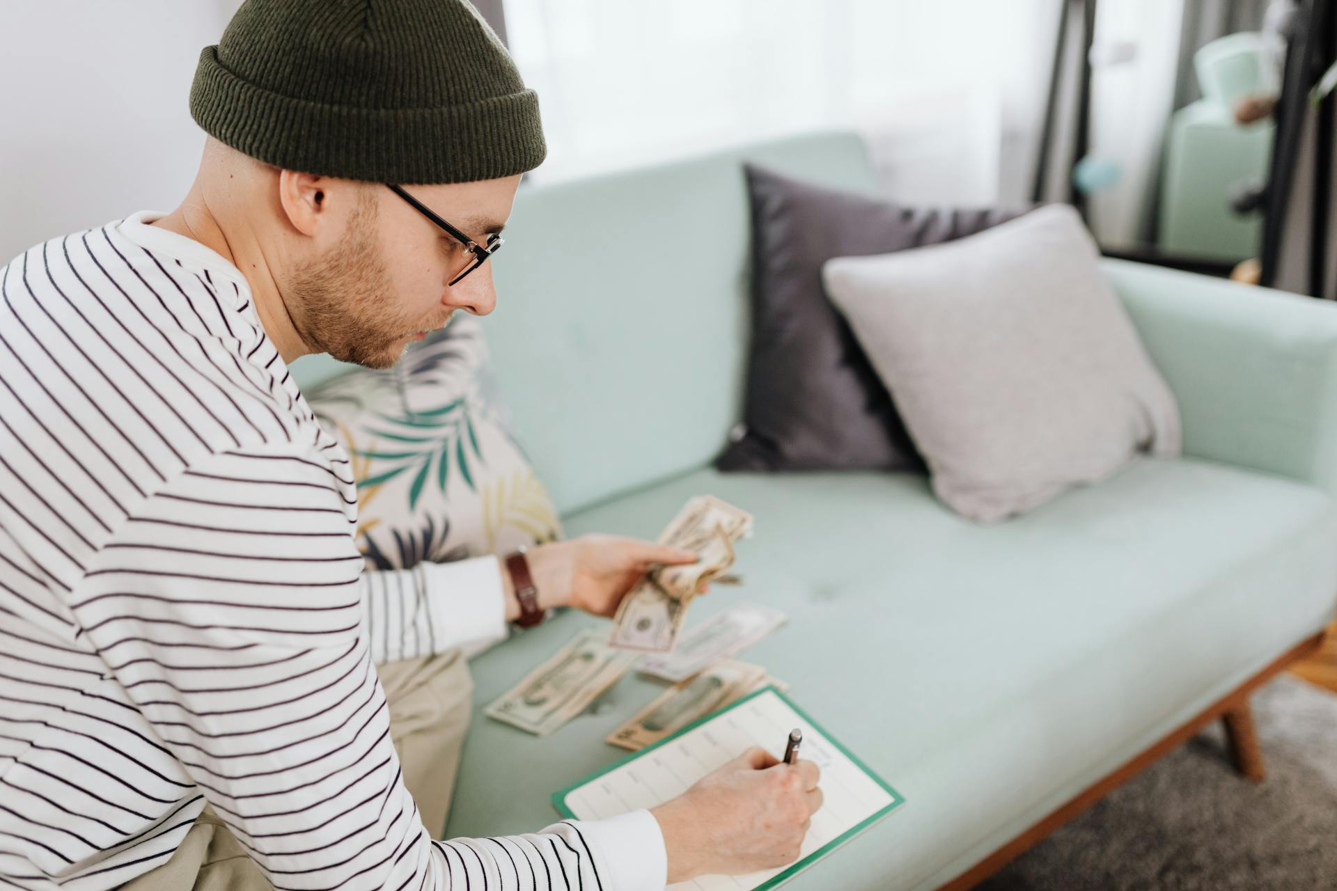 Adult man with beanie counting cash and making notes on a couch.