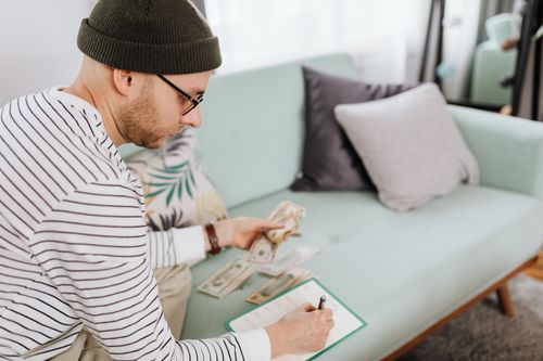 Free Adult man with beanie counting cash and making notes on a couch. Stock Photo