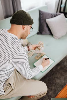 Caucasian man sitting on a couch counting cash and making notes.