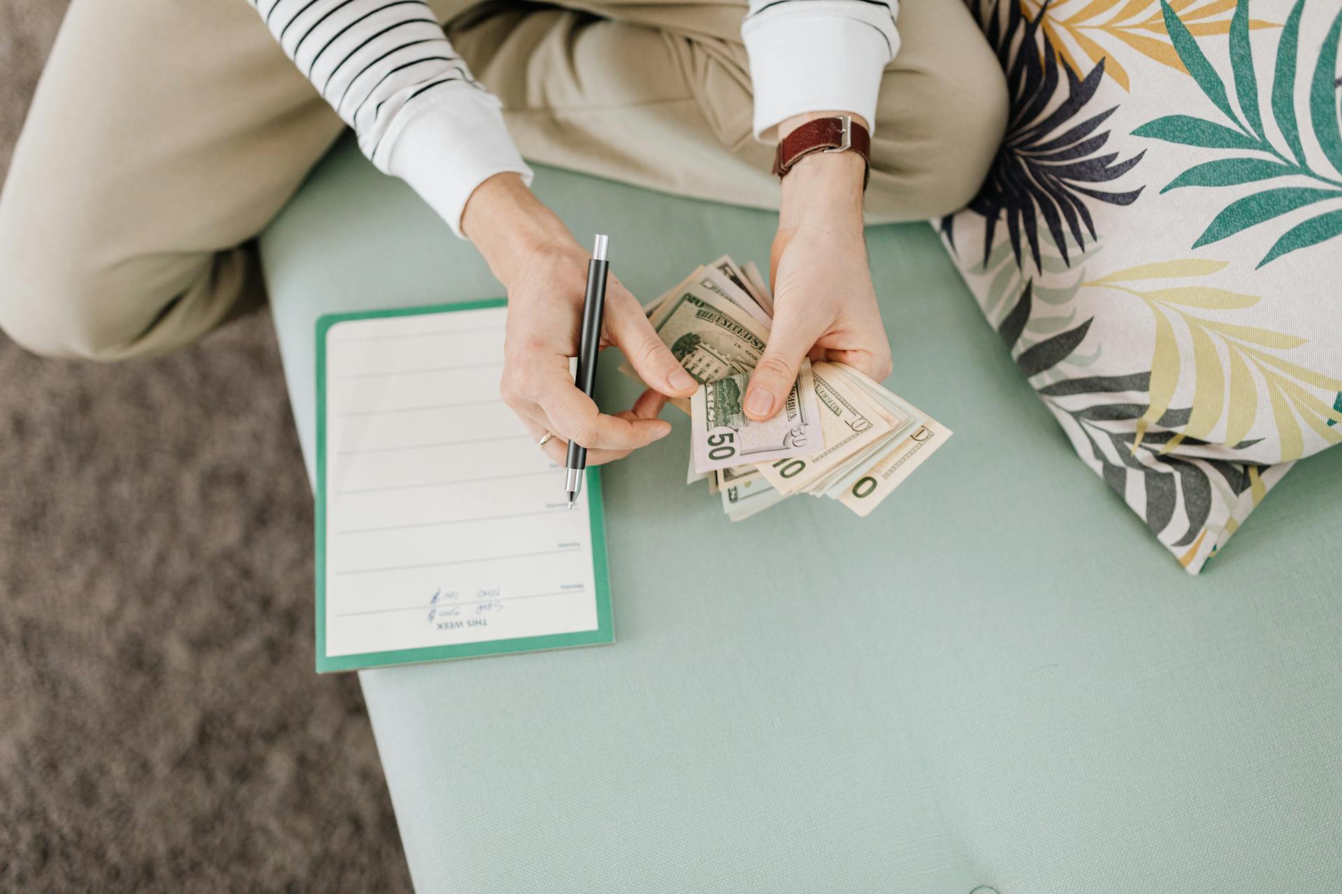 Person holding US dollar bills with a notepad and pen on a soft surface indoors.