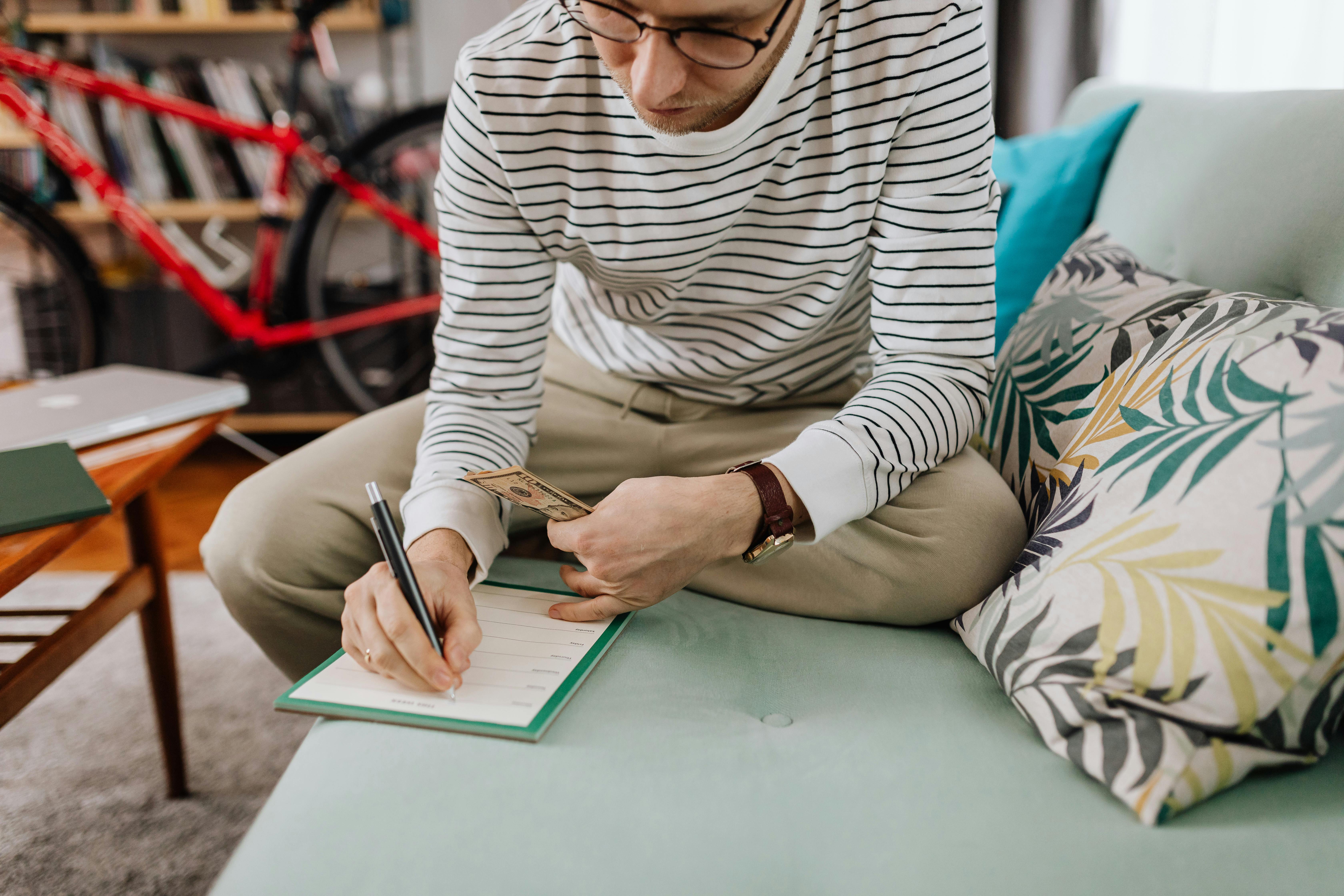 close up of a man sitting on a teal sofa holding a banknote and making notes