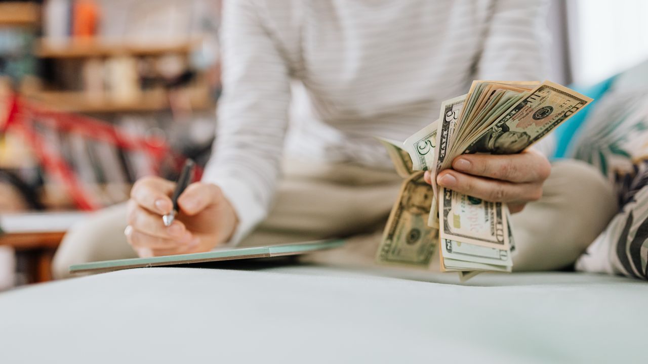 Individual counting money with pen in hand inside a cozy room setting.