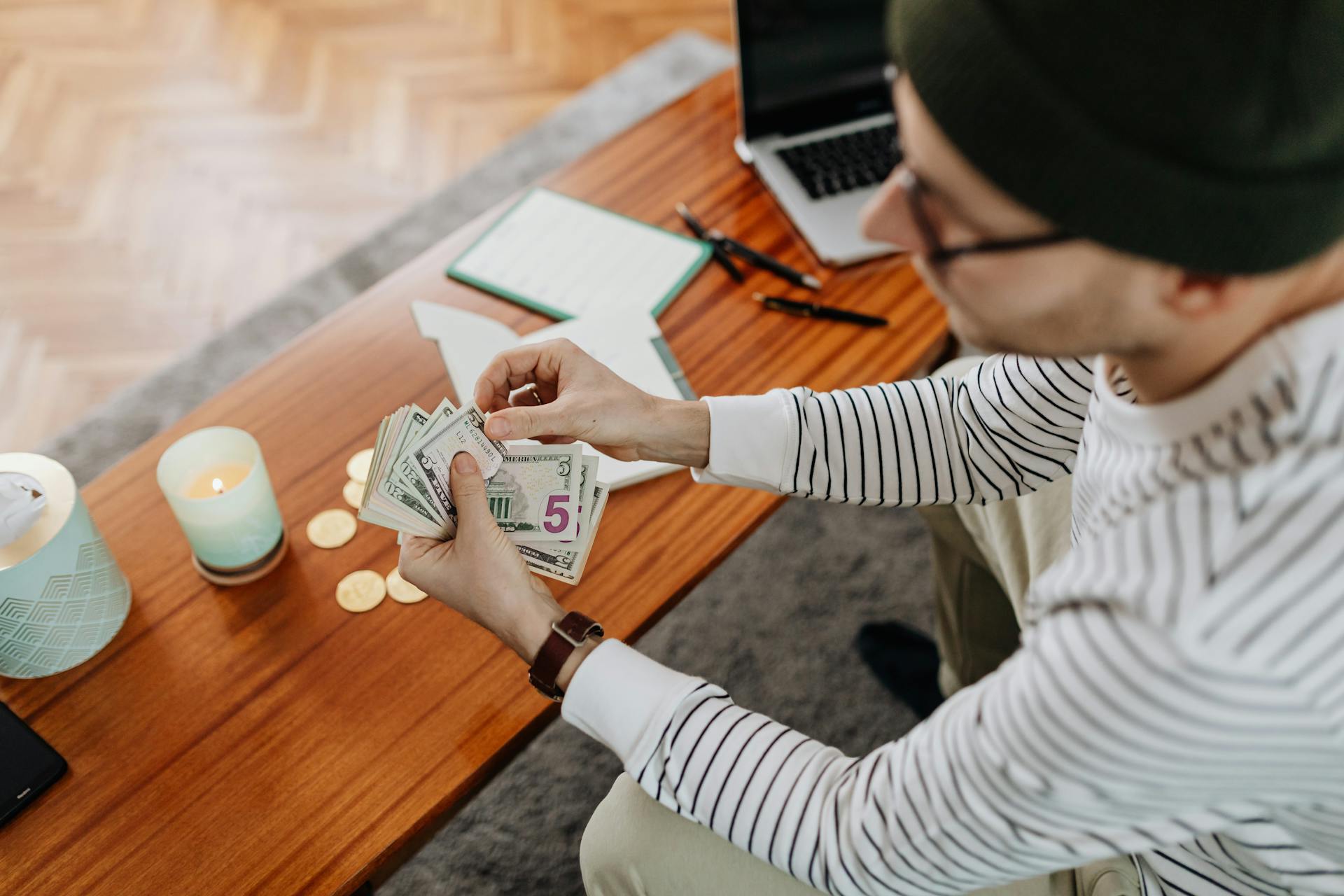 A person counts US dollars at a wooden table with a laptop, candle, and coins in view, creating a cozy atmosphere.