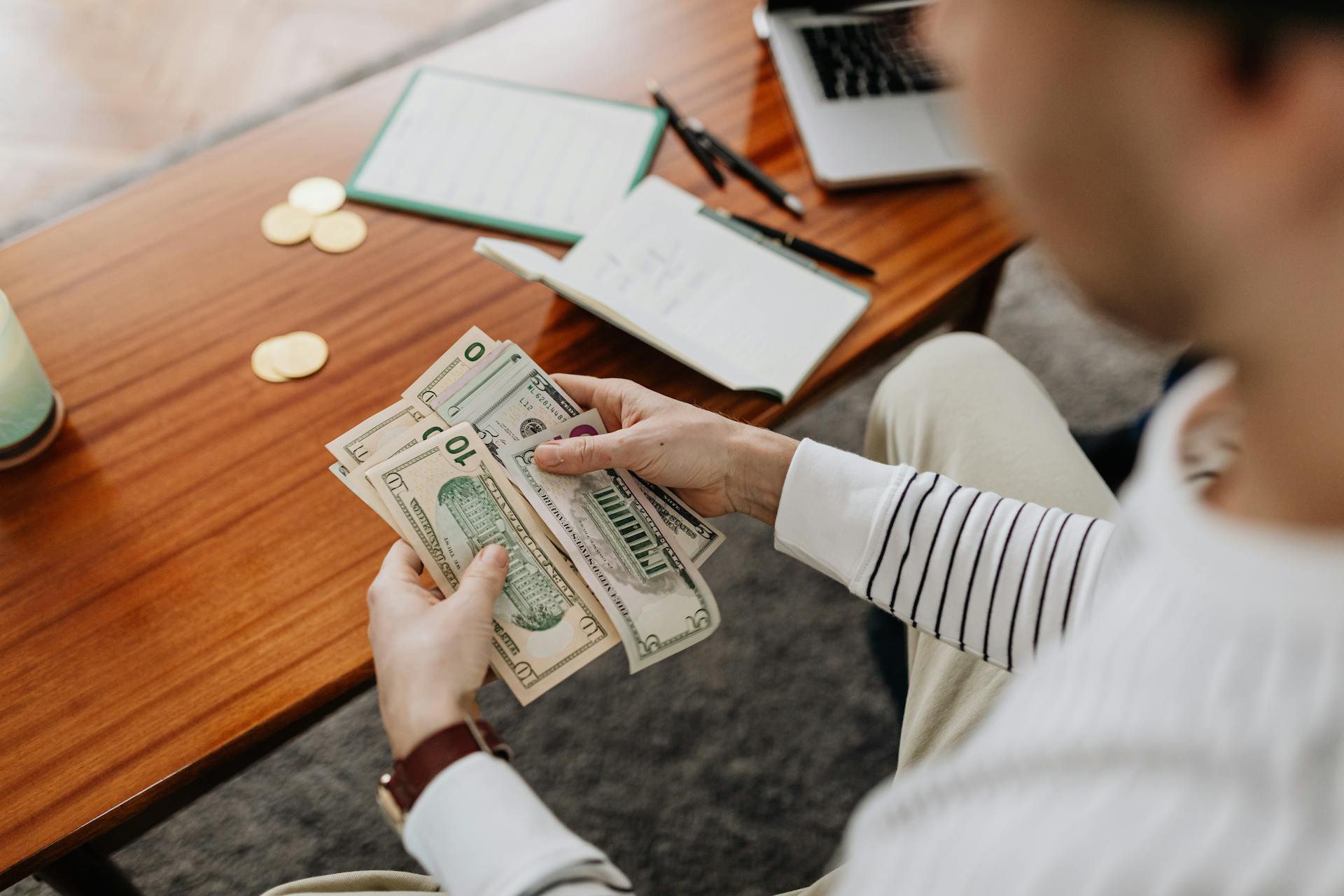 A person counts US dollar bills with a notebook and coins on the desk.