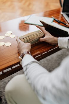 A person counting banknotes on a wooden desk indoors with coins nearby.
