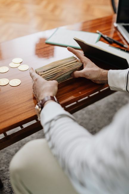 A person counting banknotes on a wooden desk indoors with coins nearby. A person counting banknotes on a wooden desk indoors with coins nearby.
