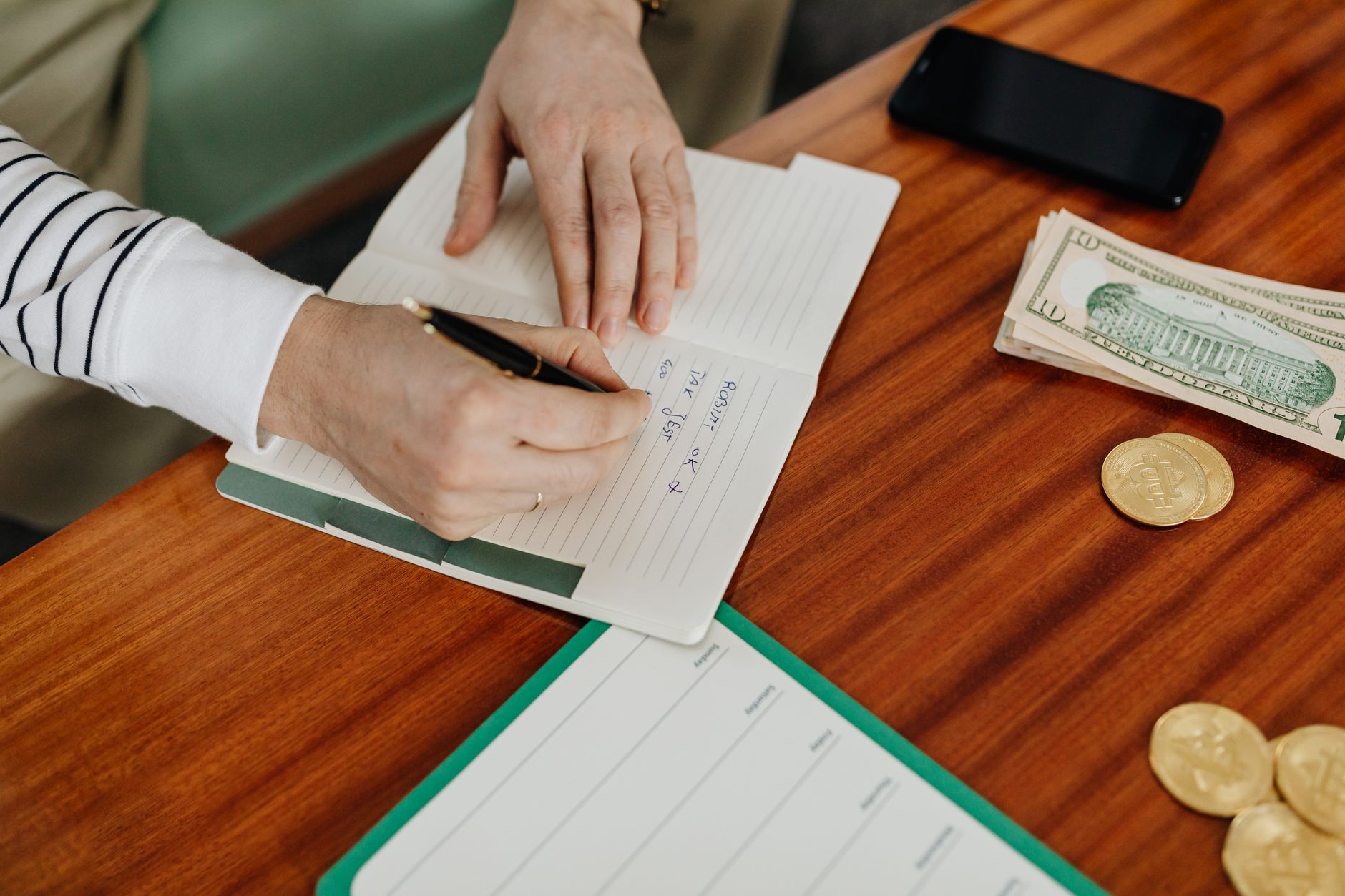Hand writing a simple budget in a notebook next to a calculator and cash.