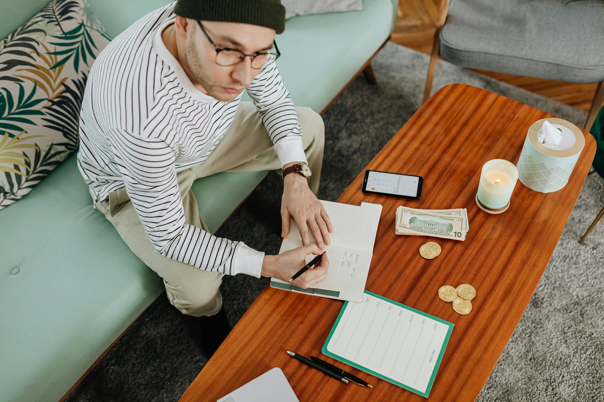 A man in a striped sweater sits on a sofa, writing in a notebook with cash and coins on a wooden table.