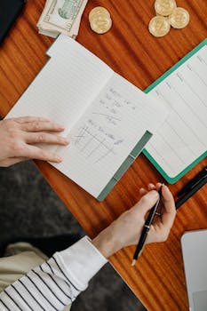 Hands holding pen and writing financial calculations in notebook with cash and coins on wooden desk.