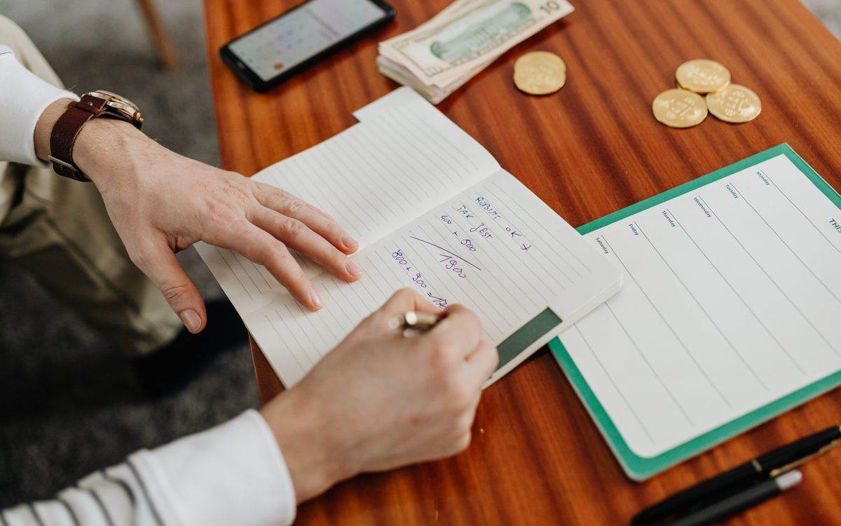 Hands writing financial calculations on notebook with money and coins on table.
