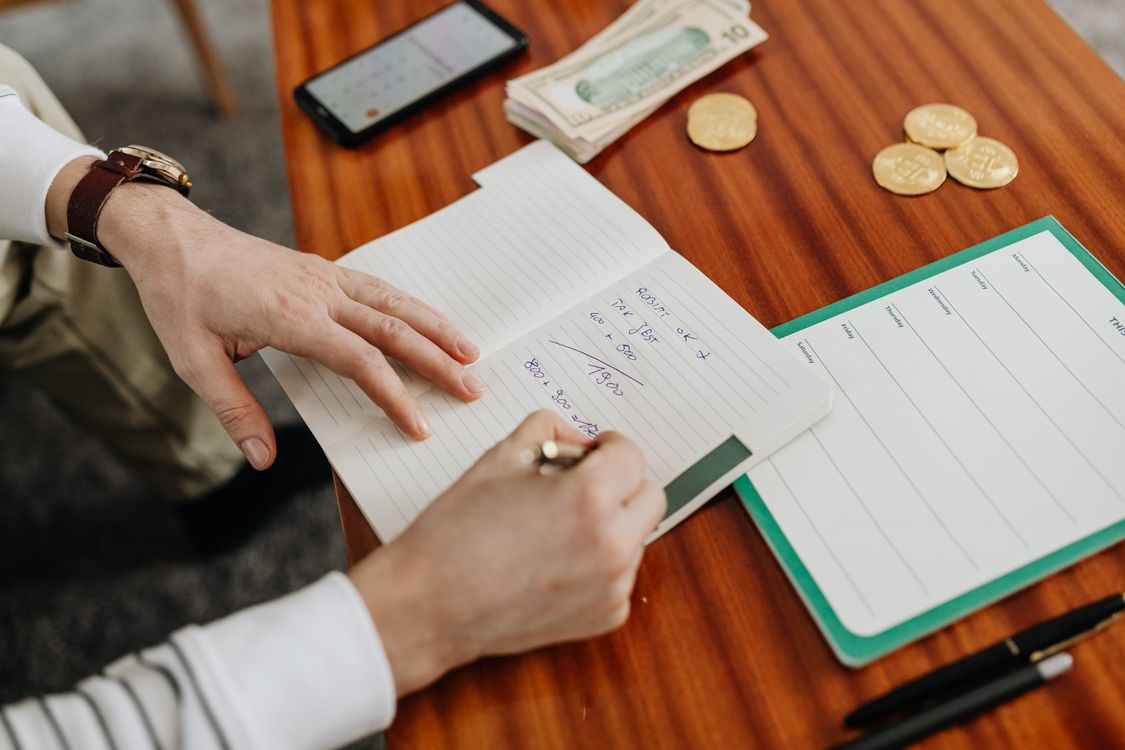 Hands writing financial calculations in a notebook beside cash and coins