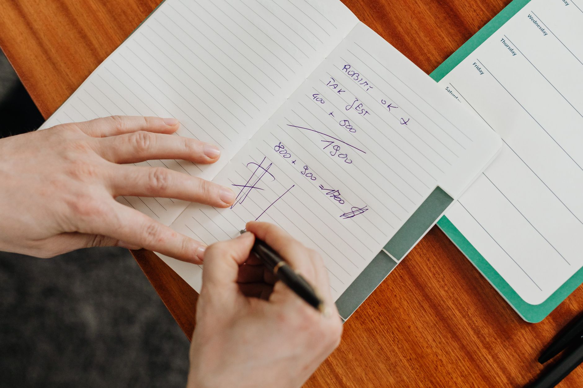 A person writing calculations in a notebook on a wooden desk.