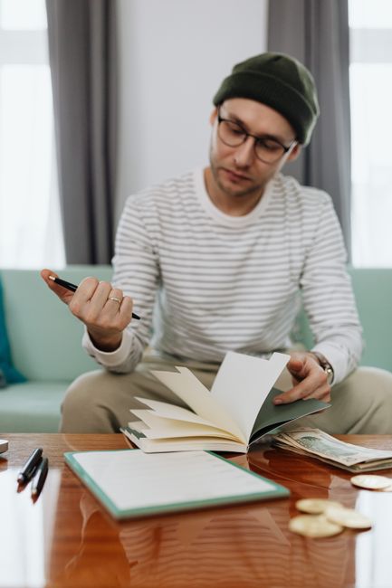 A young man wearing a beanie and glasses reviews his finances sitting on a couch. A young man wearing a beanie and glasses reviews his finances sitting on a couch.