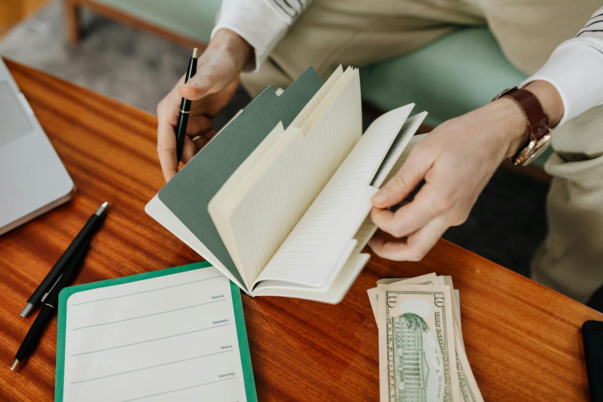 A person sits at a wooden table organizing notes in a notebook and handling cash.