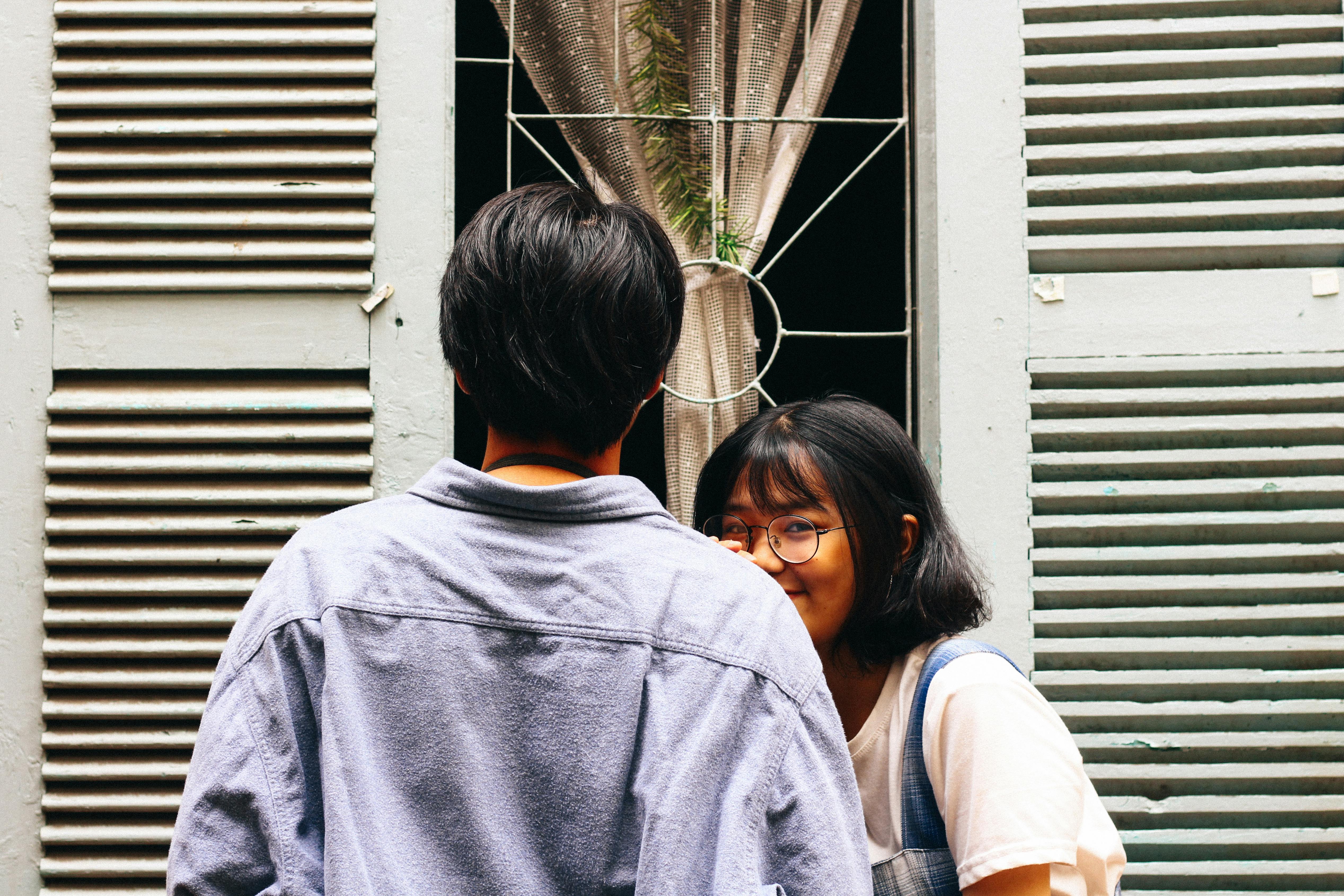 Young cheerful couple in casual clothes standing near window and hugging while smiling
