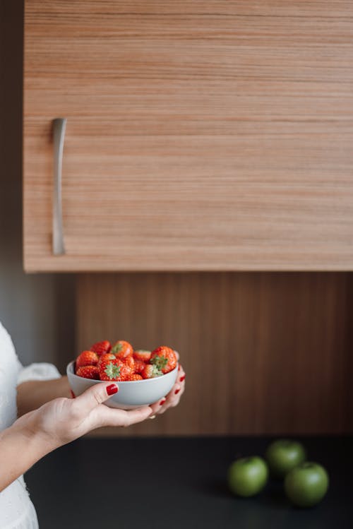 Unrecognizable female with bowl of strawberry standing near counter with green apples