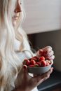 Side view of crop pensive young female with long wavy blond hair enjoying taste of sweet ripe strawberries during breakfast in kitchen