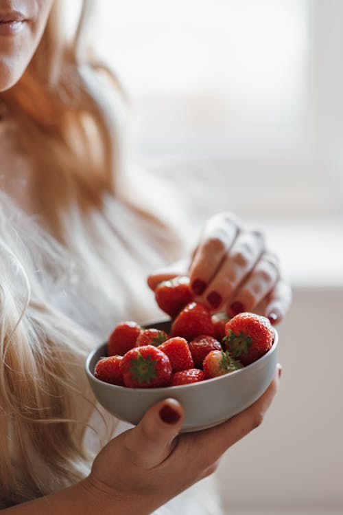 High angle of crop unrecognizable female with long blond hair eating fresh red tasty strawberries while standing at home on sunny day