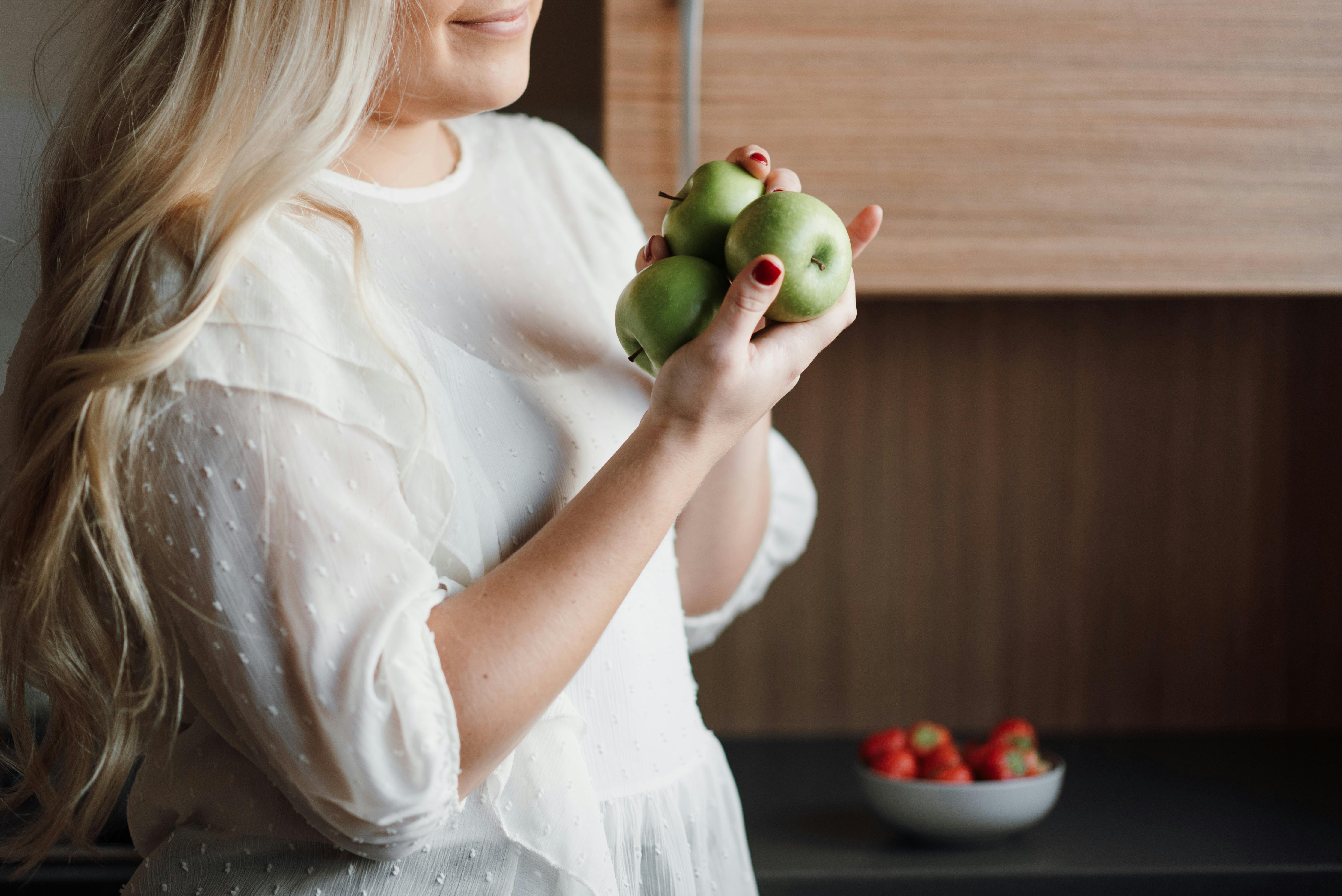 Side view of crop unrecognizable positive lady with long blond hair smiling while holding fresh green apples in hands standing near table with bowl of ripe strawberries