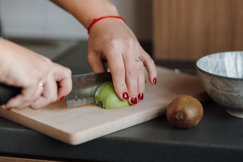 Crop anonymous female cutting ripe green apple on wooden cutting board in kitchen in daytime