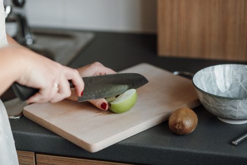Free Crop unrecognizable female cutting green apple with sharp knife while standing in light kitchen near counter with kiwi and bowl near cutting board Stock Photo